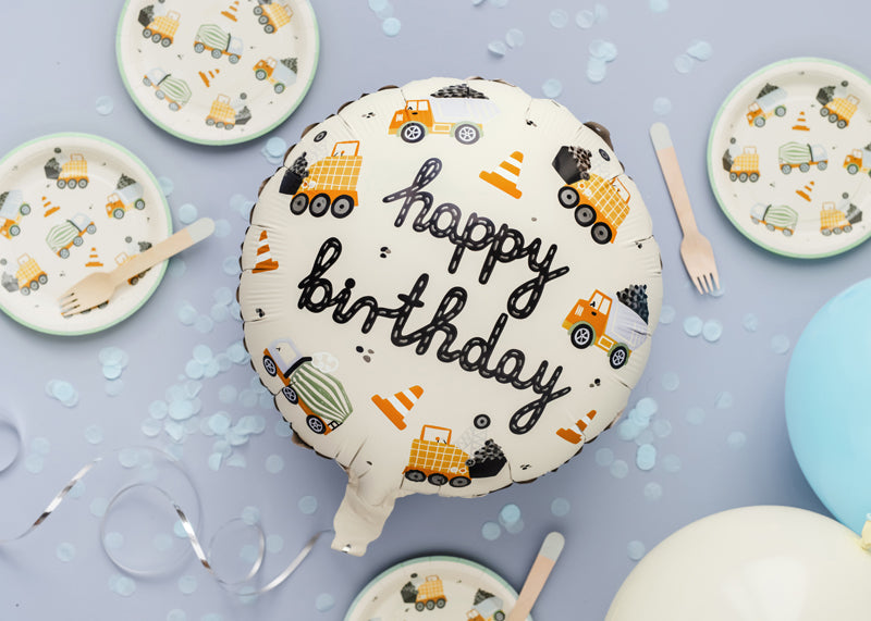 Birthday balloon with 'happy birthday' text and construction vehicle pattern on a table with plates and utensils.
