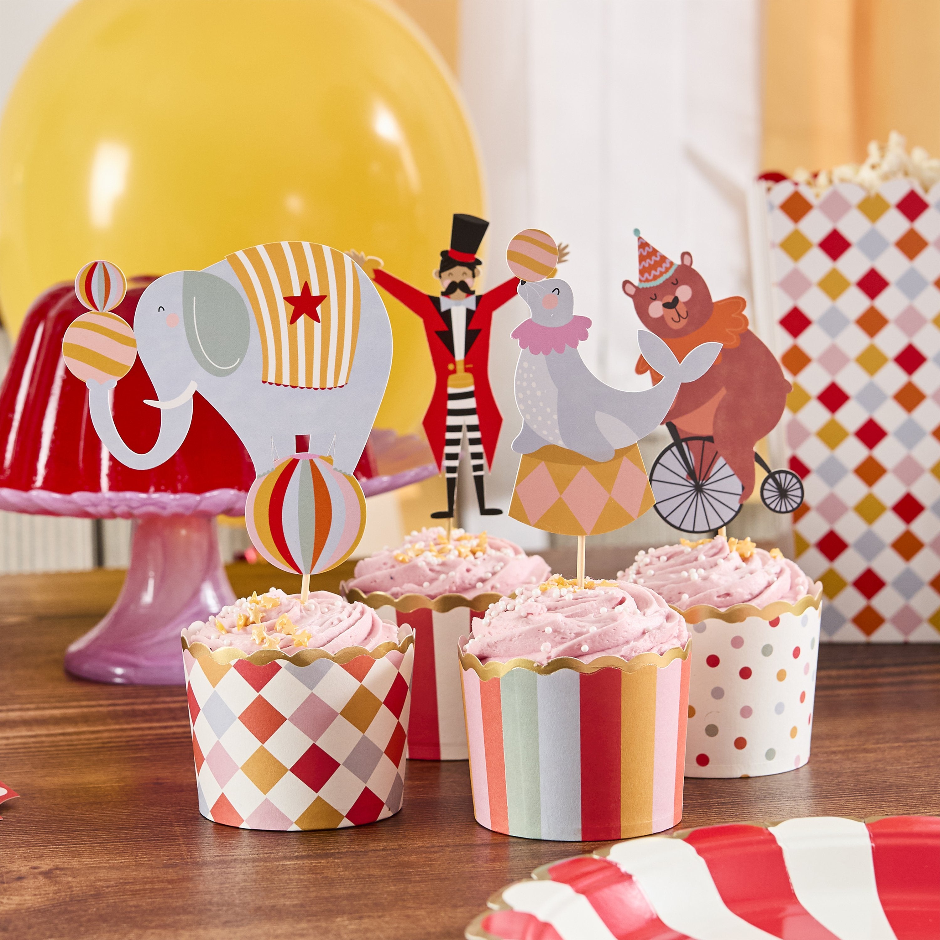 A set of circus-themed party cupcake toppers with various colorful designs, including an elephant, a clown, and a horse, displayed on a table with striped and polka dot patterned toppers.