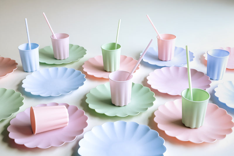 Assorted frill plastic plates in shades of pink, purple, green, and blue, with designs resembling a scallop or frill, displayed with disposable cups on top.